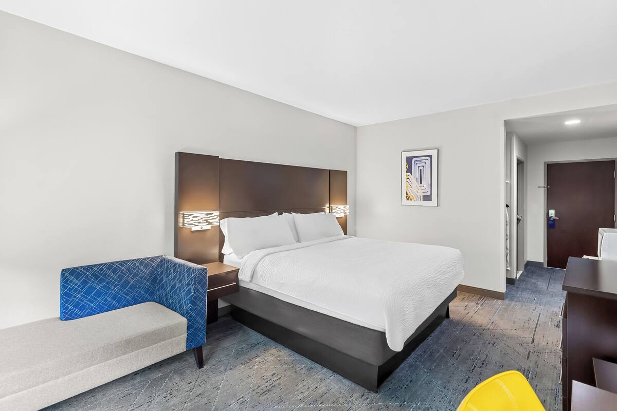 A modern hotel room with a large bed, a blue accent chair, a desk, and a small yellow chair.
