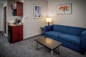 A compact hotel room with a kitchenette, a blue sofa, a small table, and two wall-mounted artworks.