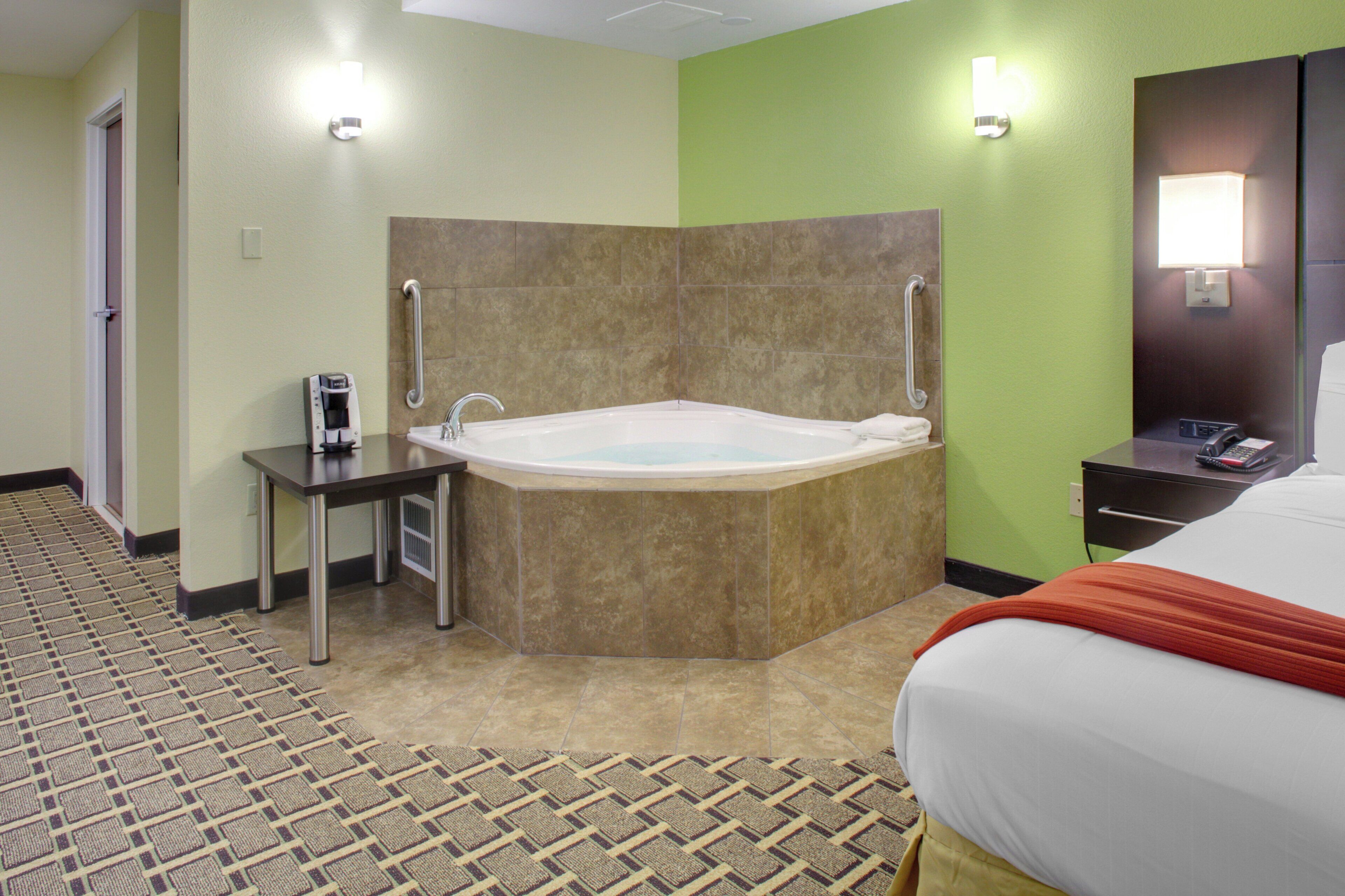 A hotel room with a large bathtub, a small table with a coffee maker, a bed with a red blanket, and a bathroom with a towel rack.