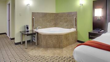 A hotel room with a large bathtub, a small table with a coffee maker, a bed with a red blanket, and a bathroom with a towel rack.