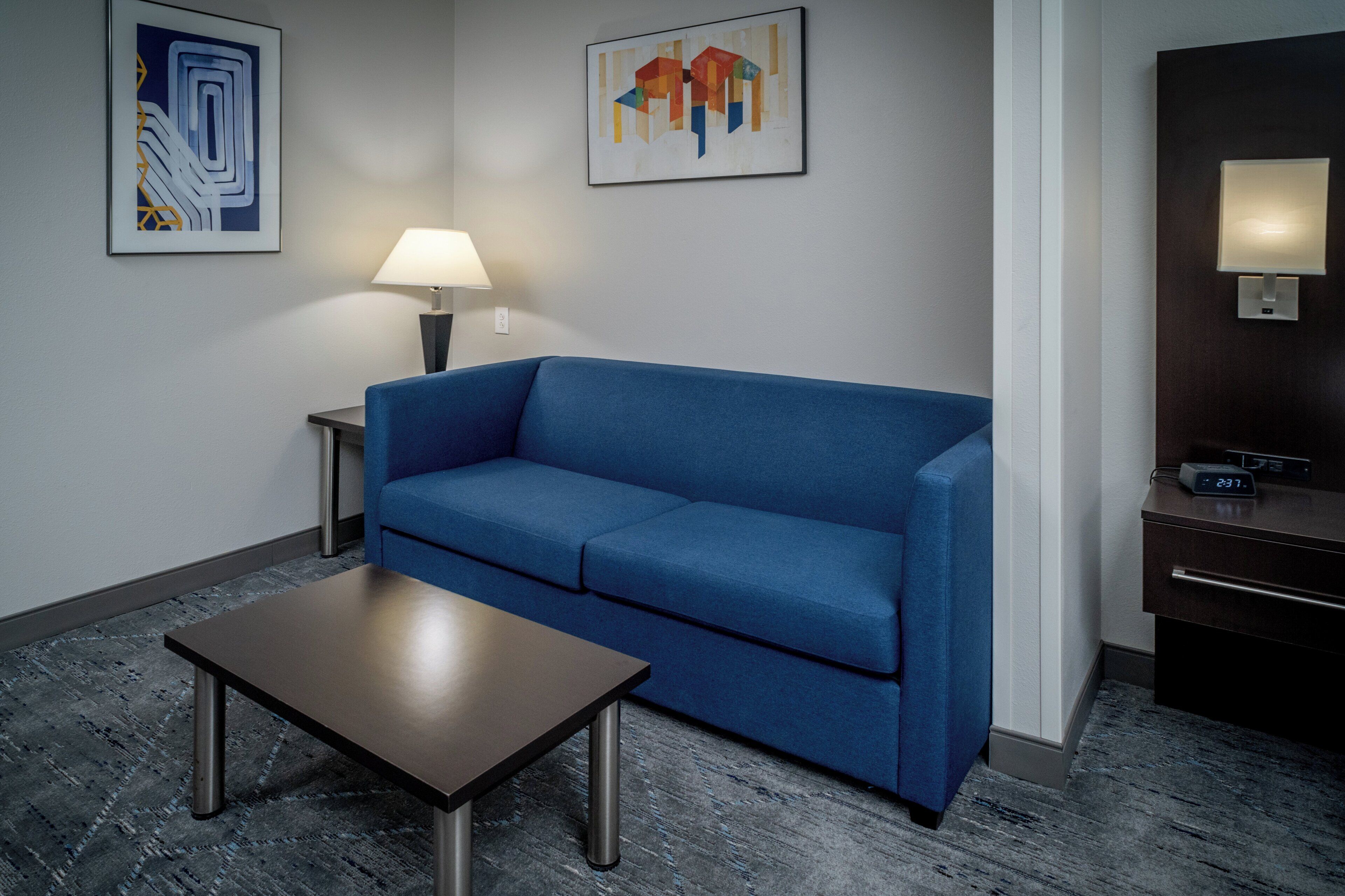 A hotel room with a blue sofa, a small table, a lamp, and artwork on the walls.