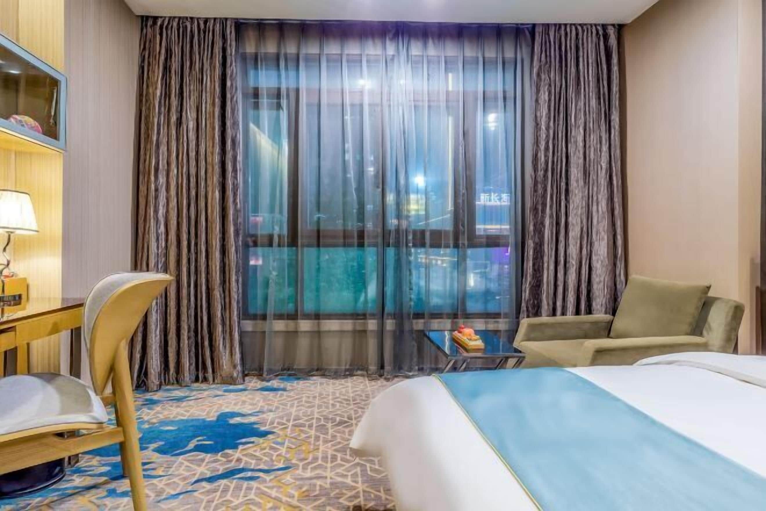 A hotel room with a bed, a desk, a chair, a small table with a fruit bowl, and a large window with curtains.