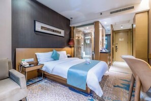 Ett hotellrum med en stor säng, sängbord, ett skrivbord, en stol, en inramad tavla på väggen och ett badrum som syns genom en glasdörr.