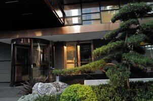 Una entrada de edificio moderna con puerta de vidrio, un jardín bien cuidado con arbustos variados y una gran formación rocosa.