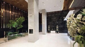 Un vestíbulo amplio con diseño moderno, que presenta una columna central, un muro de mármol negro y una barandilla de vidrio con una pequeña fuente.