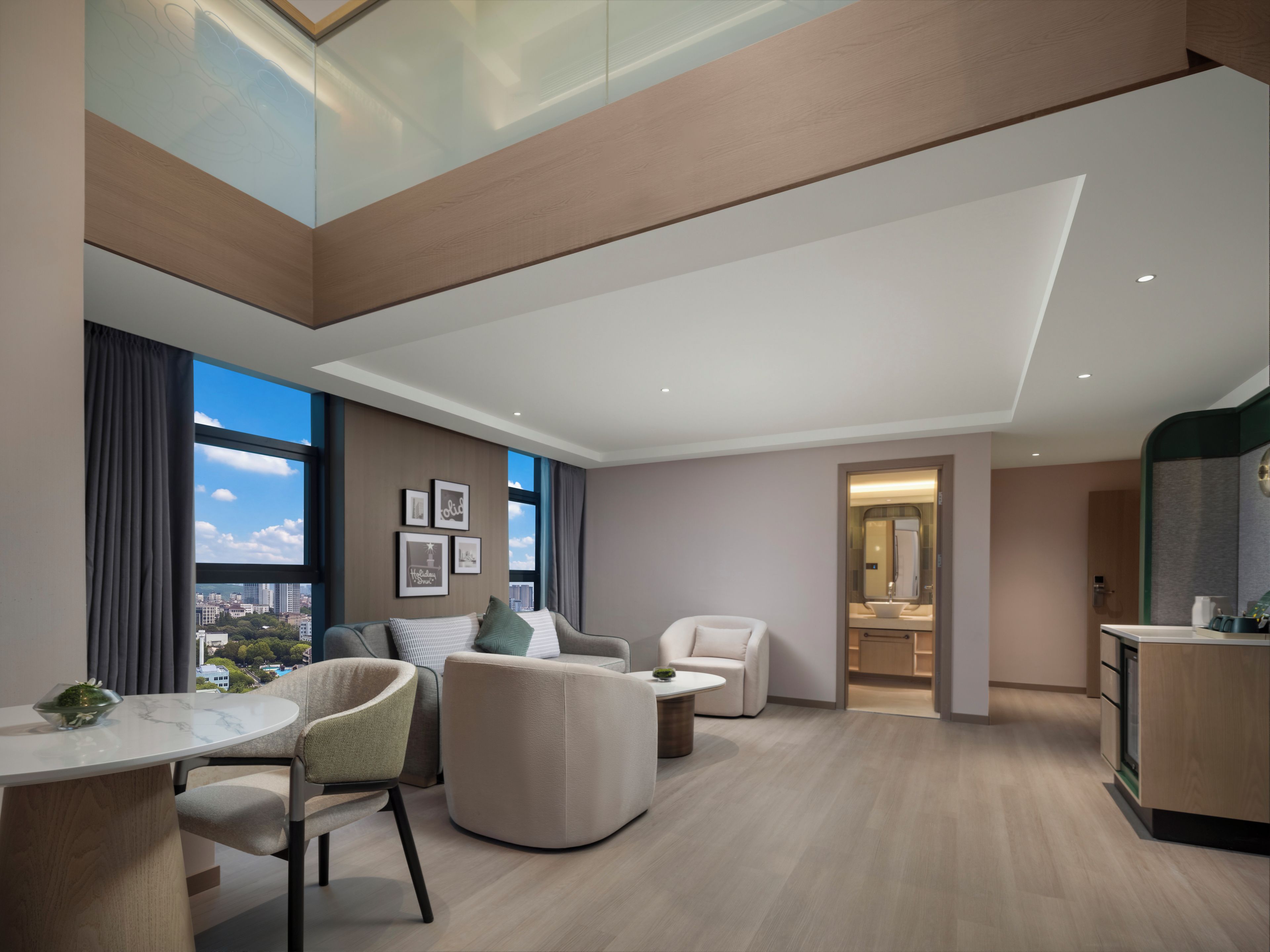 A modern hotel room with a dining area, a seating area with a sofa and chairs, a kitchen, and a large window offering a city view.