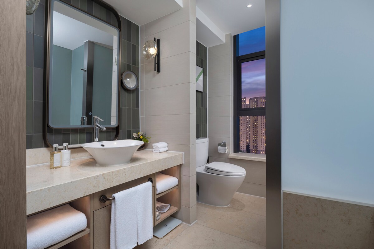 A modern bathroom with a large mirror, a sink, a toilet, and a window with a city view.