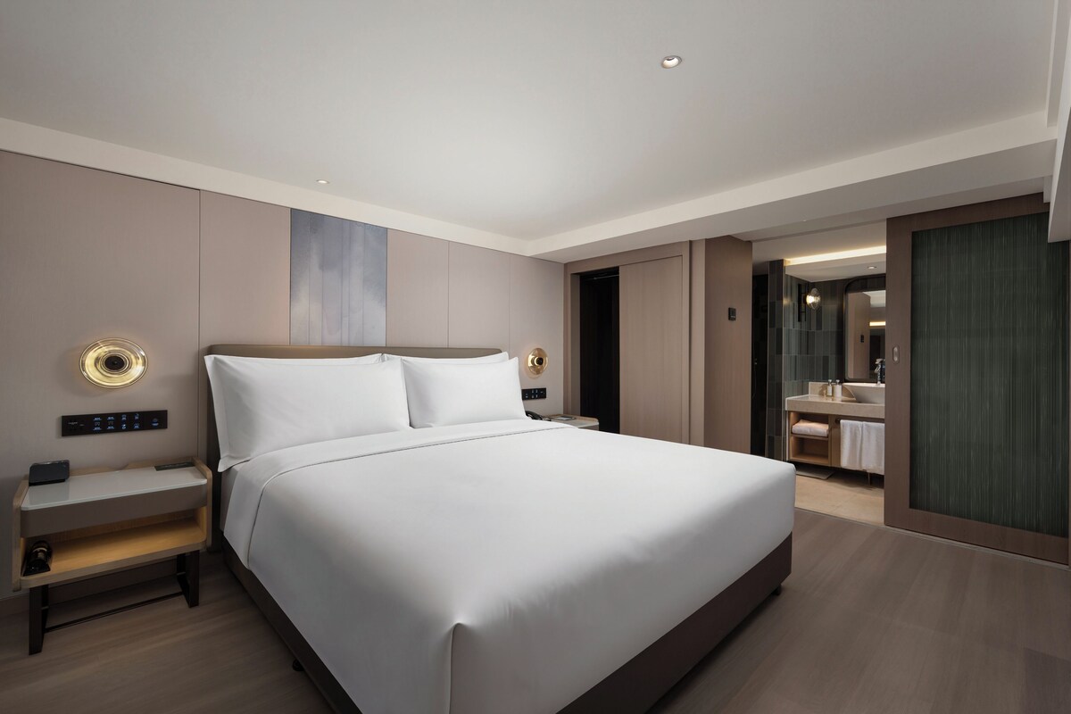 A modern hotel room with a large bed, bedside tables, a nightstand, and a bathroom with a sink and mirror.