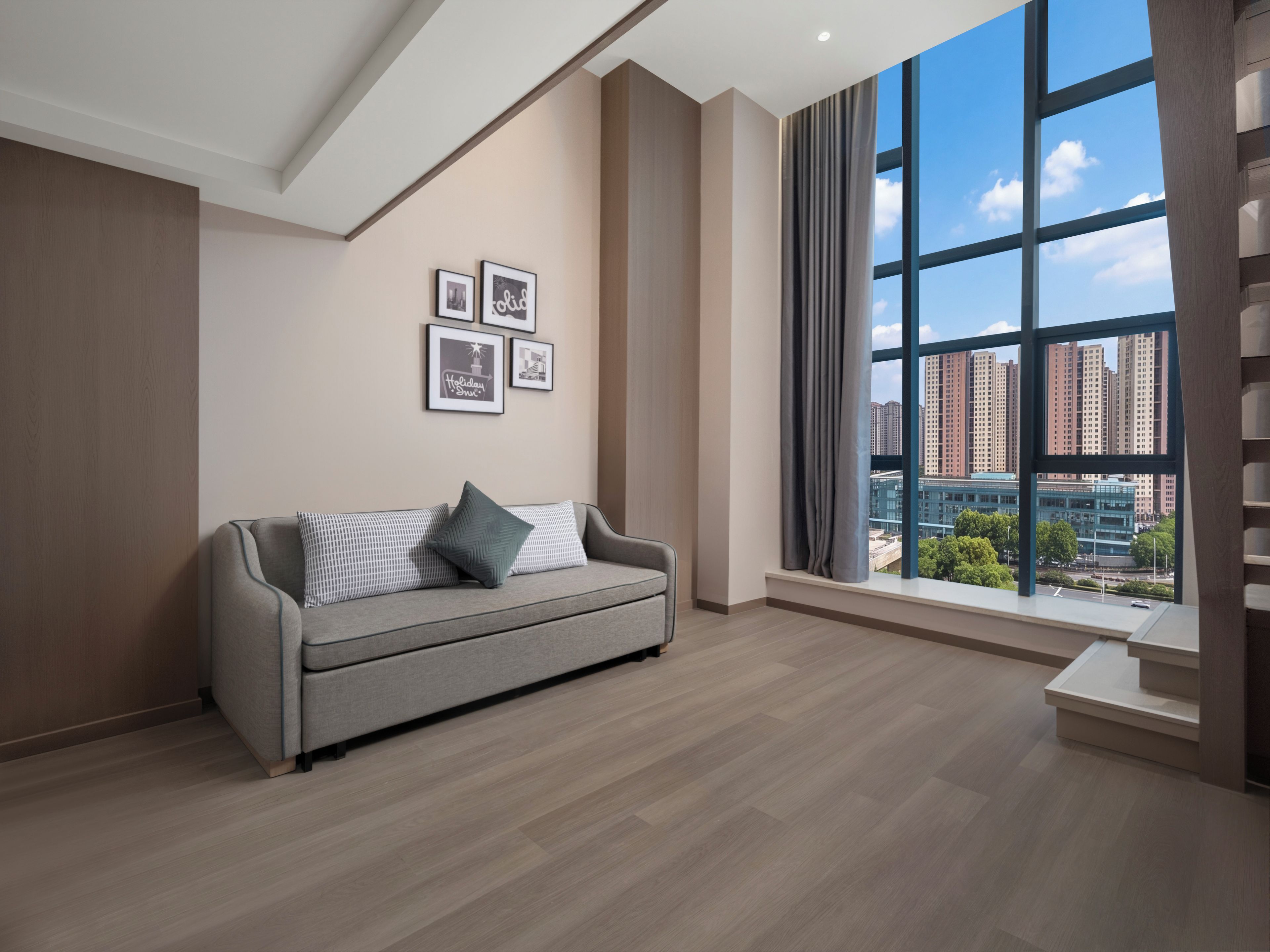 A modern living room with a grey sofa, a large window with a city view, and framed pictures on the wall.