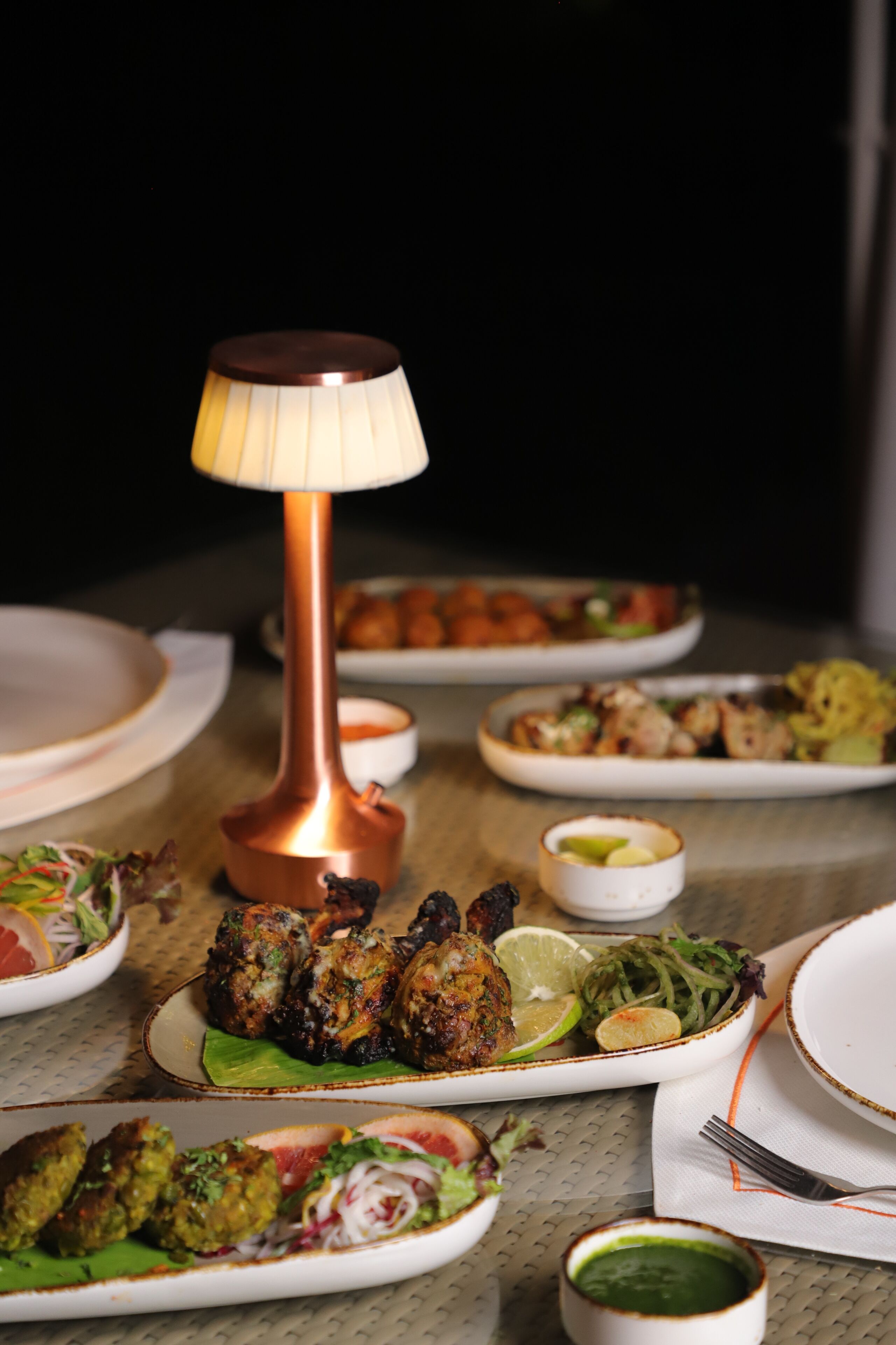 Uma mesa de jantar com diversos pratos, uma luminária cor de cobre e uma pequena tigela de molho.