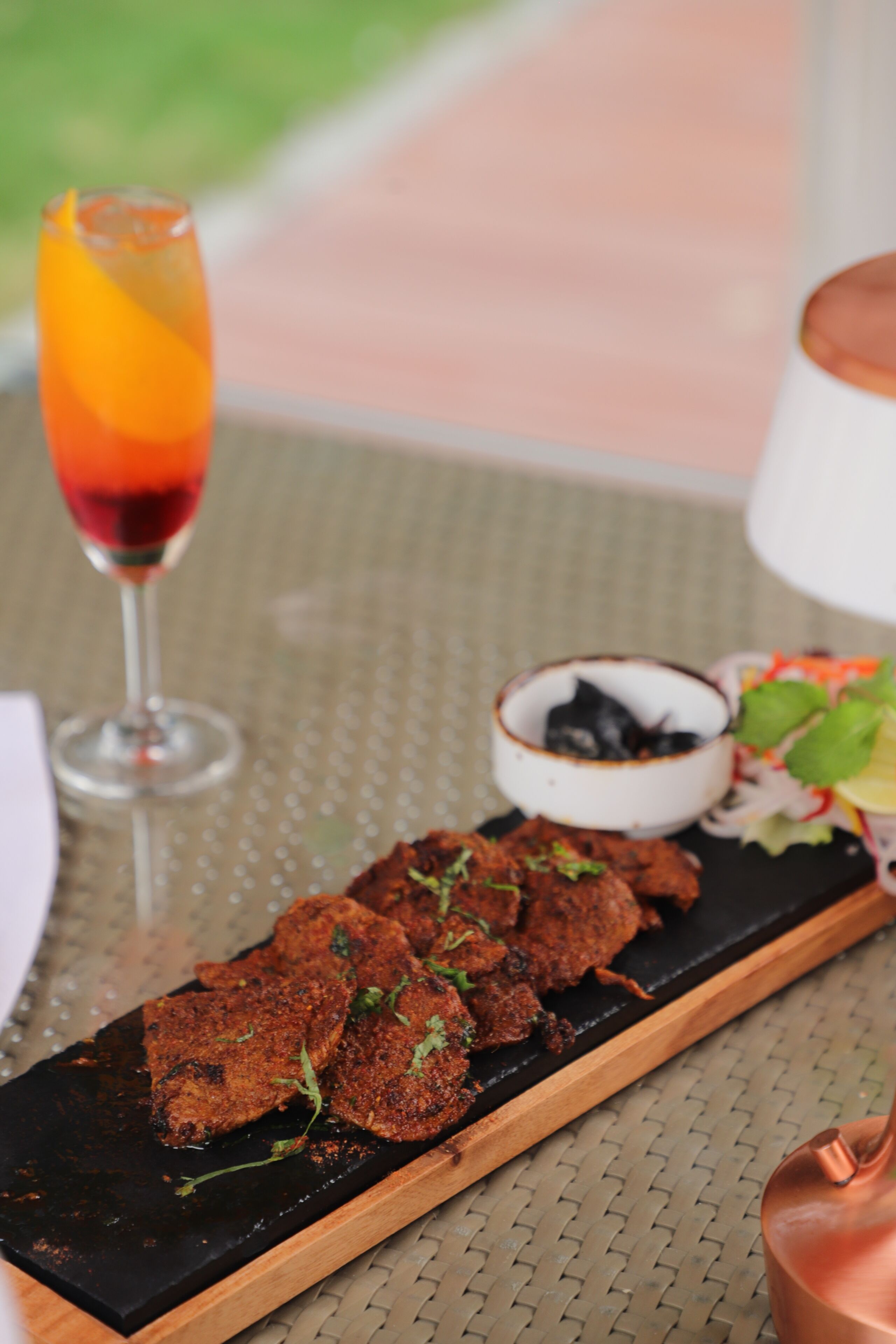A wooden serving board with grilled meat, a glass of red wine, a bowl of olives, and a salad on a woven tablecloth.