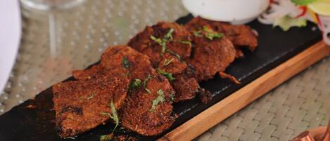 A wooden serving board with grilled meat, a glass of red wine, a bowl of olives, and a salad on a woven tablecloth.