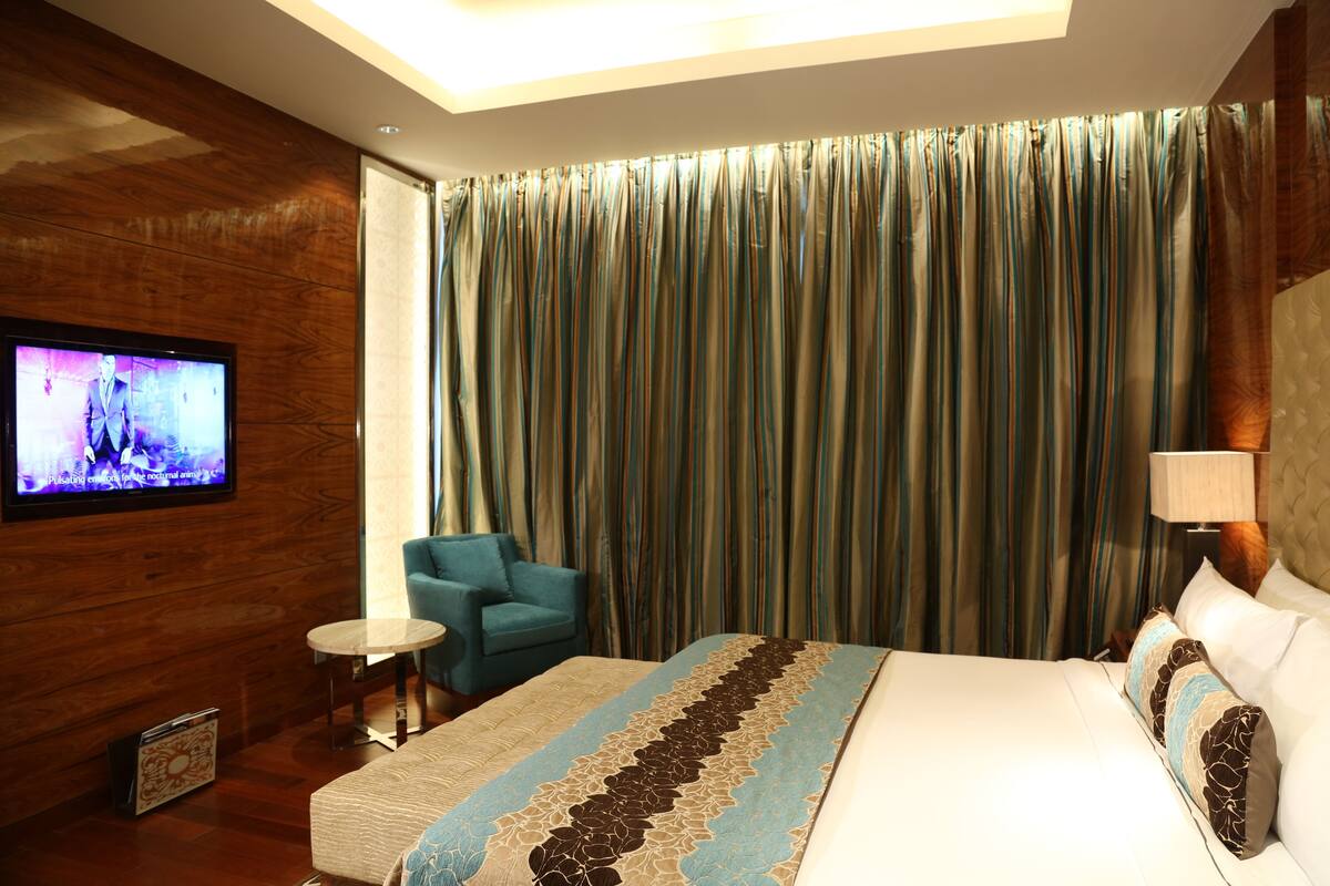 A hotel room with a large bed, a television on the wall, a blue armchair, a small table, and a lamp.