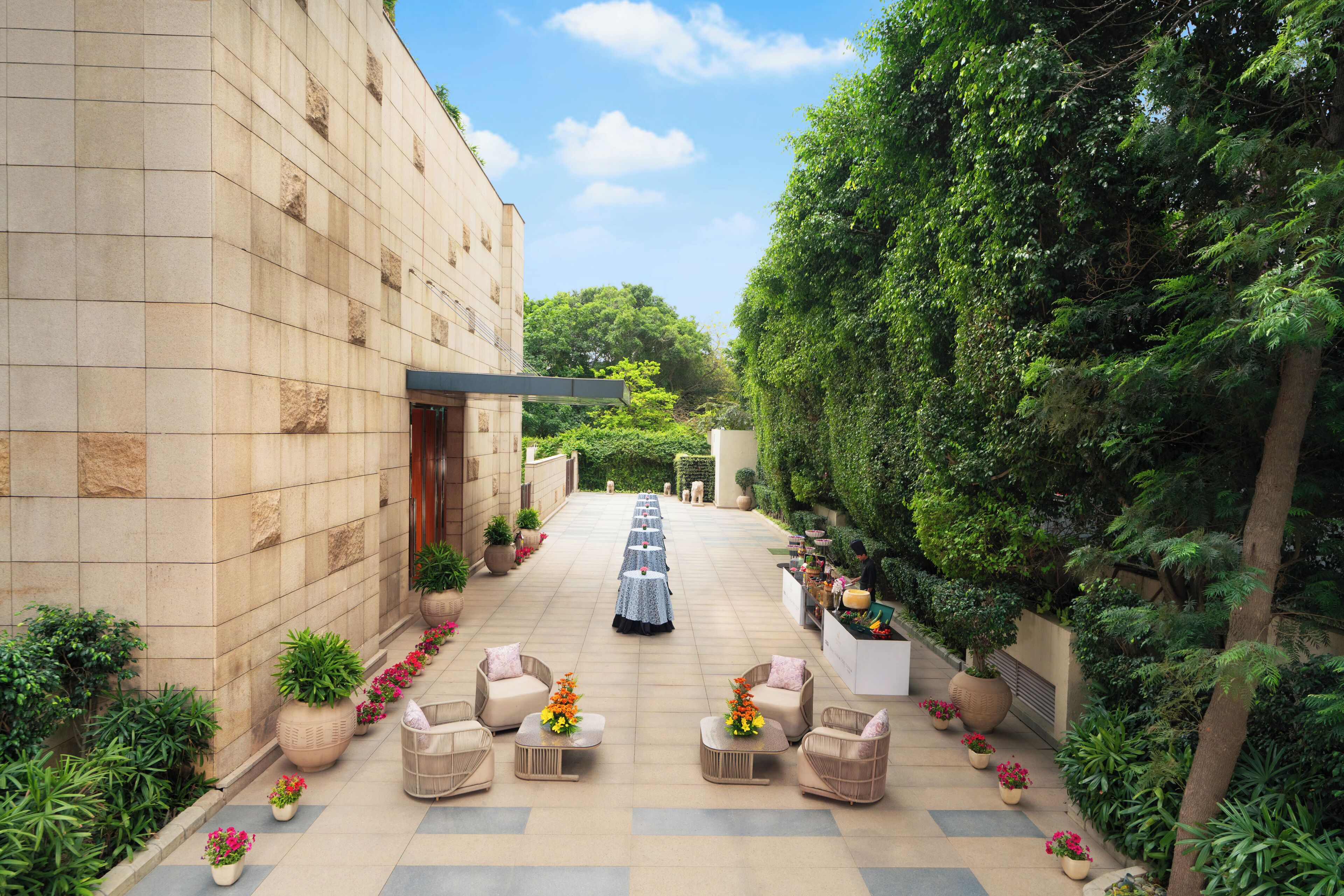 A courtyard with a row of tables and chairs, a pathway leading to a building entrance, and lush greenery on the sides.