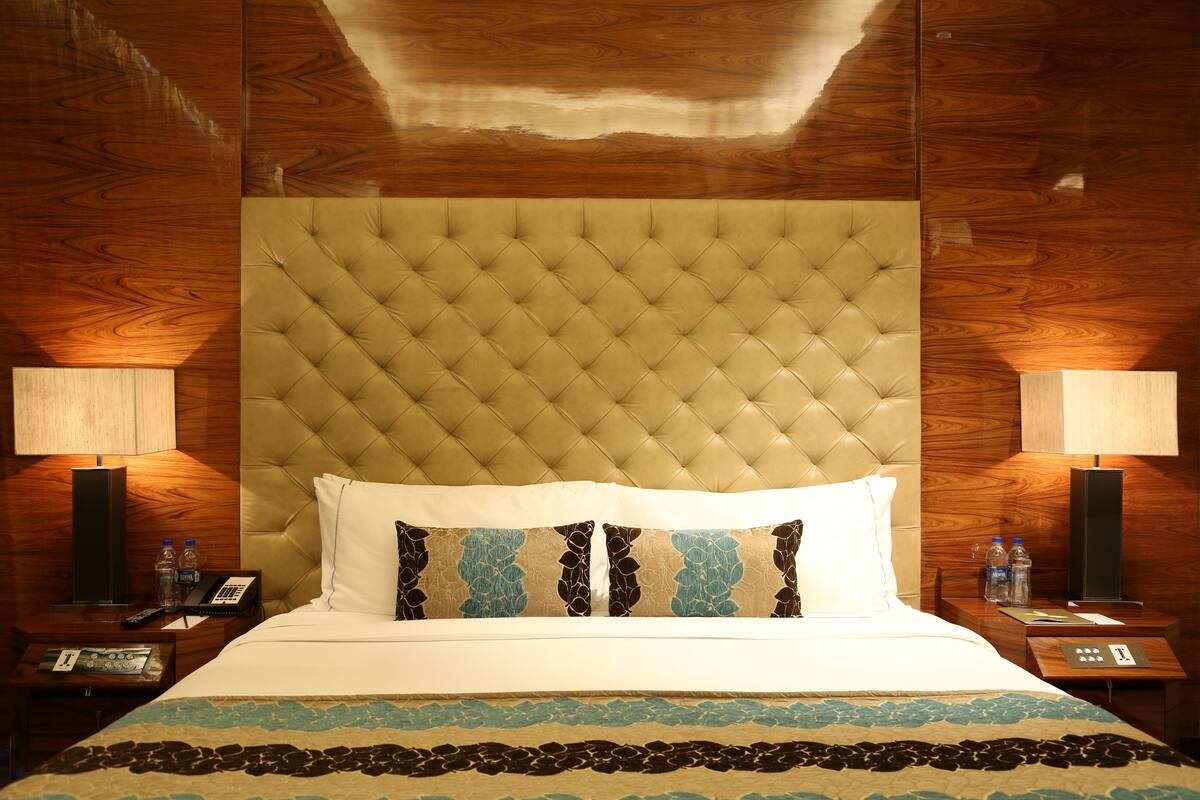 A hotel room with a tufted headboard, a bed with white and patterned pillows, a bedside table with a lamp, and a nightstand with a phone and water bottles.