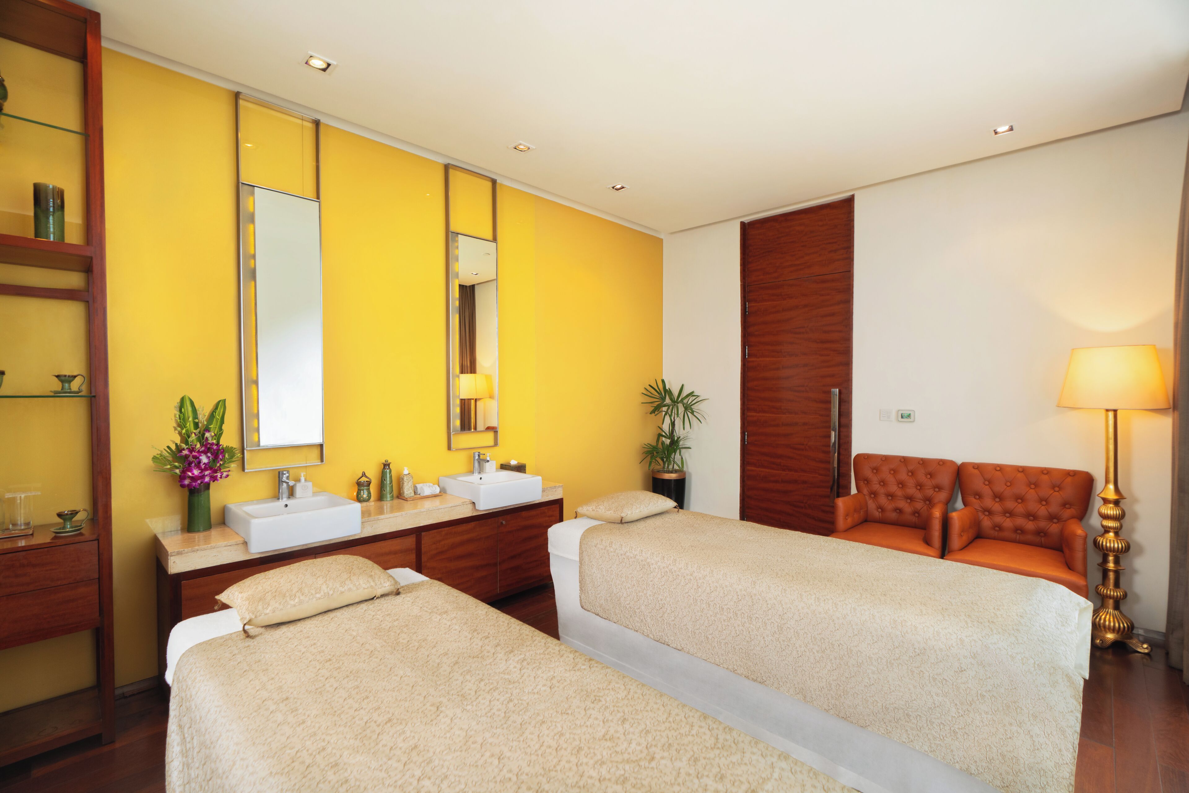 A hotel room with two beds, a large mirror, a wooden wardrobe, a bathroom with a sink, and a bedside table with a lamp.