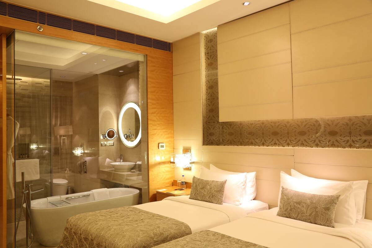 A hotel room with a large bathtub, a separate shower area, a round mirror, a bedside table with a lamp, and a bed with two pillows.