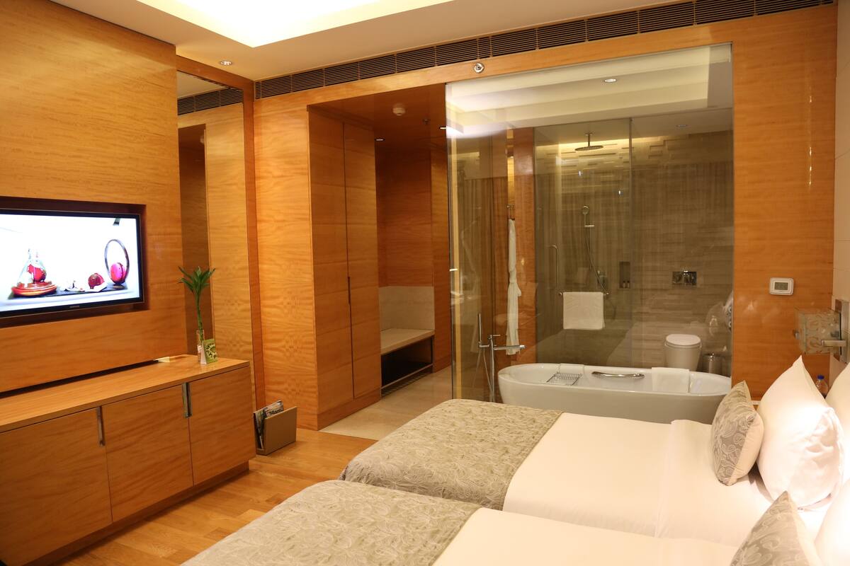 A modern hotel room with a wooden finish, a TV mounted on the wall, a glass-enclosed shower area, and a large bed with pillows.