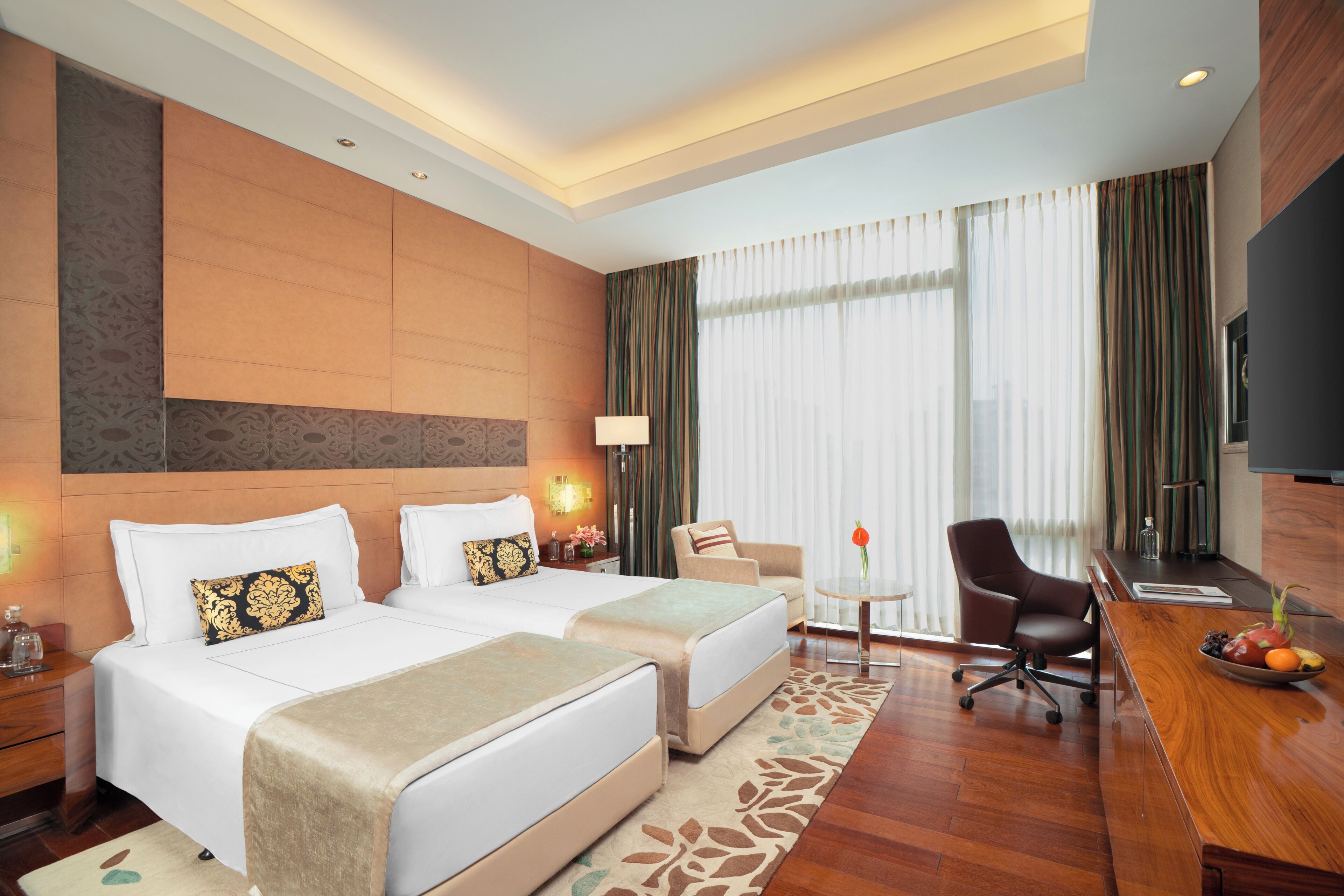 A hotel room with two beds, a desk, a chair, a TV, and a large window with curtains.