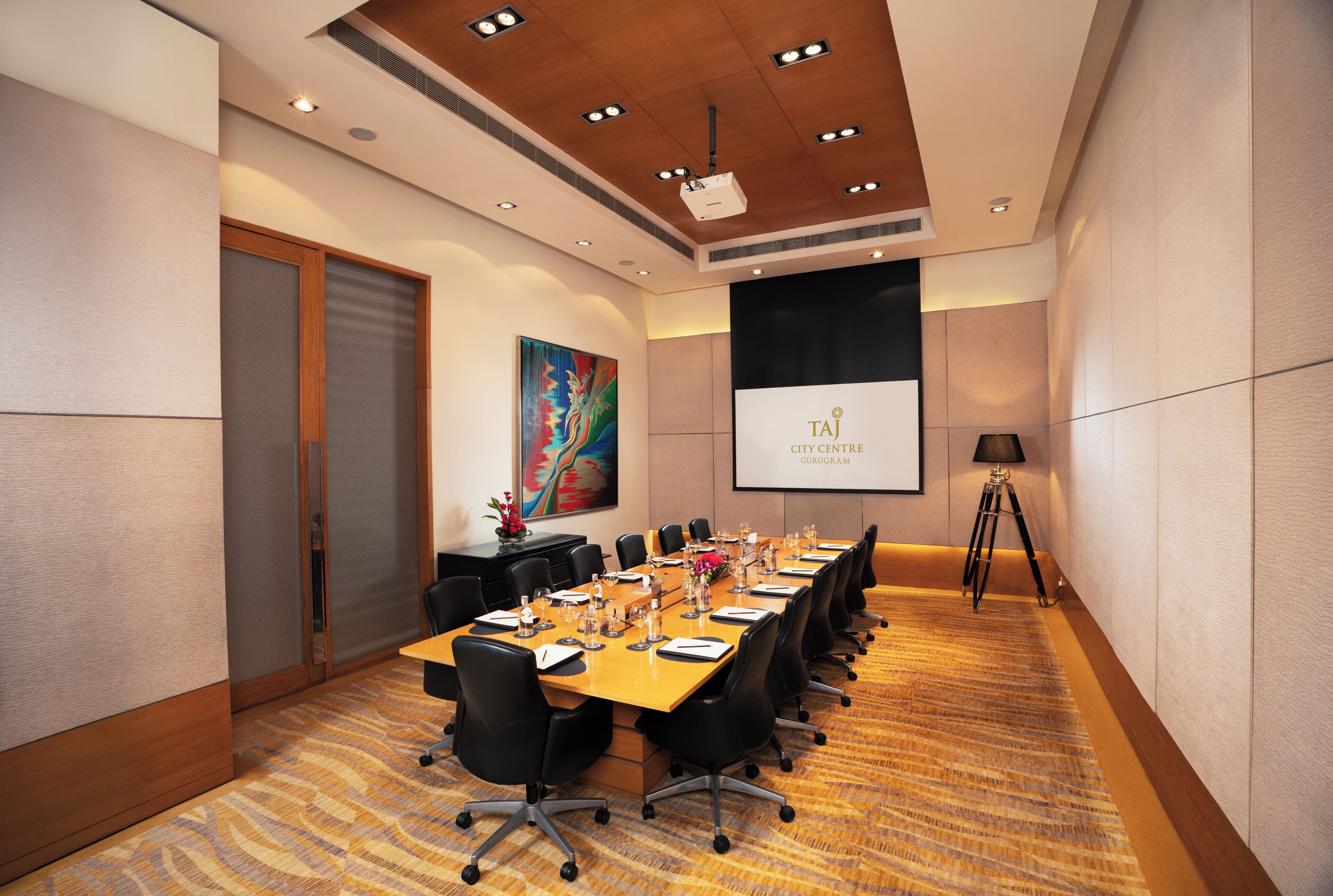 A conference room with a long table, chairs, a projector screen, and a painting on the wall.