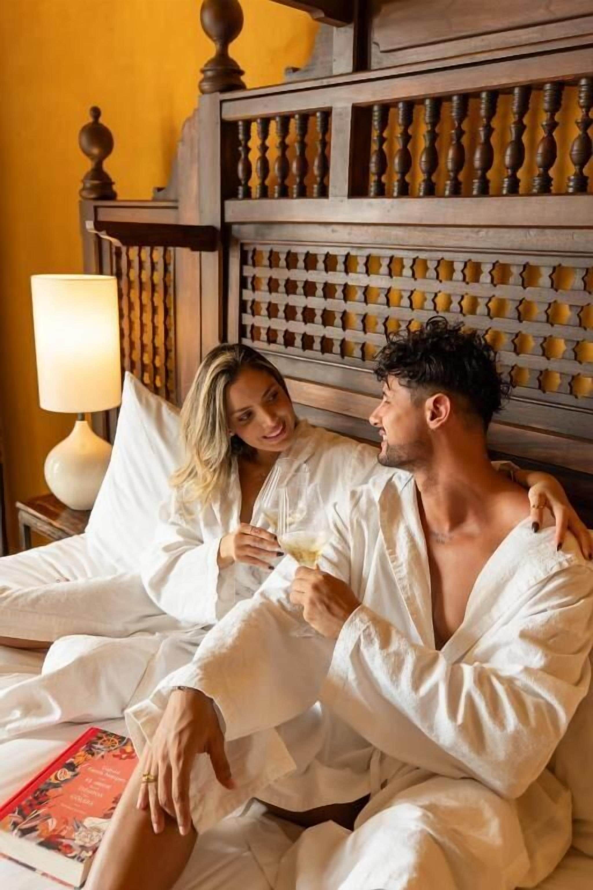 A couple in white bathrobes on a bed with a wooden headboard and a bedside lamp.