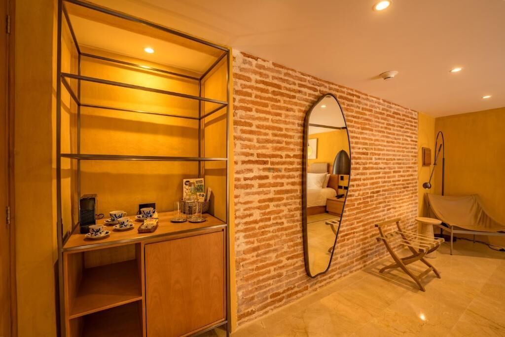 A room with a brick wall, a large mirror, a wooden bench, and a shelf with cups and a coffee maker.
