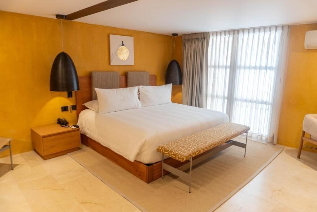 Un dormitorio con cama, mesitas de noche, un banco, una lámpara colgante, un cuadro en la pared y una ventana con cortinas.