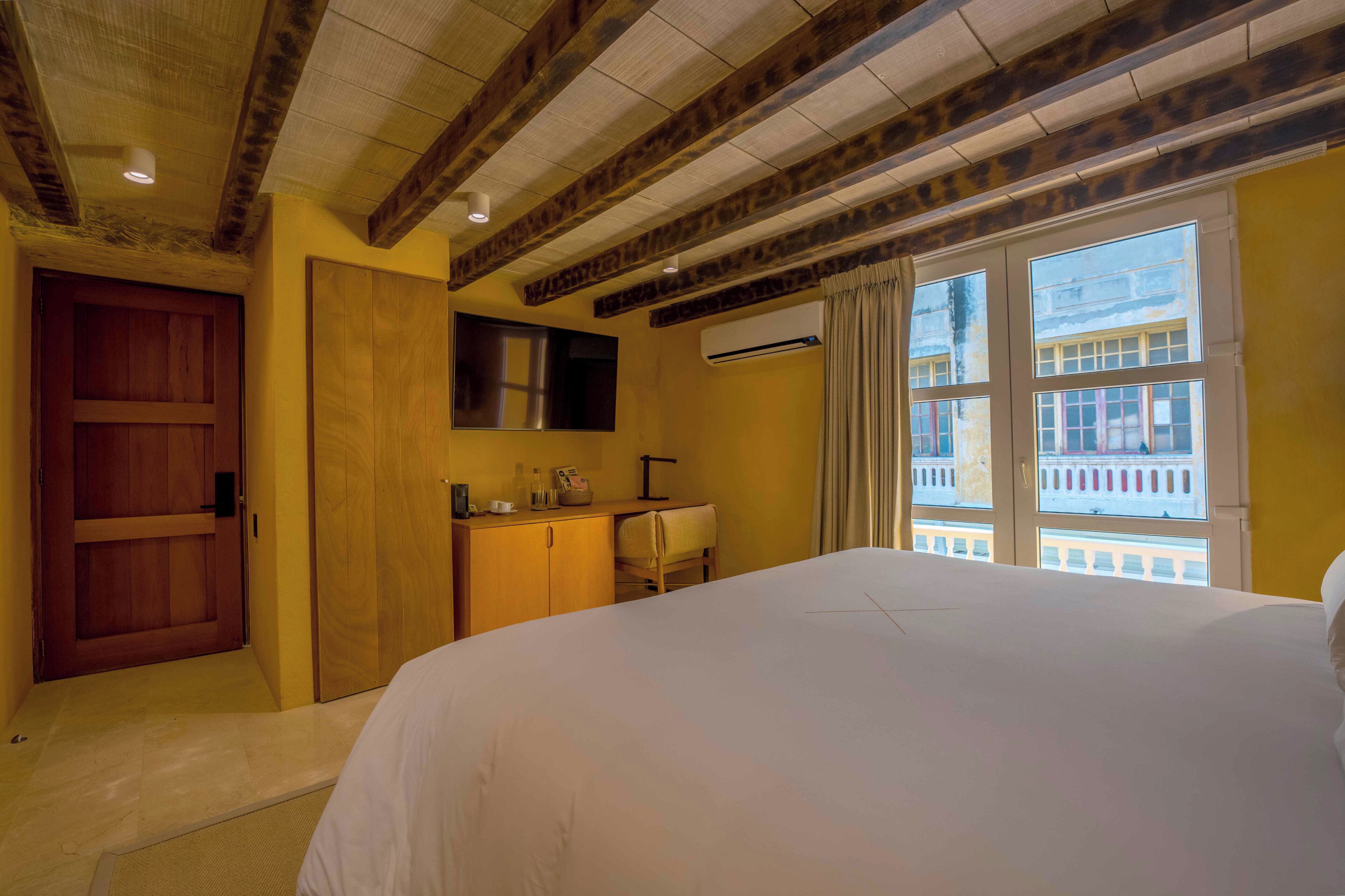 Habitación de hotel con una cama grande, vigas de madera en el techo, un escritorio con una silla, una ventana con cortinas y un equipo de aire acondicionado.