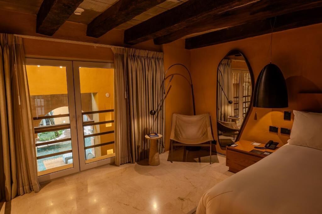 Une chambre d’hôtel équipée d’un lit, d’un bureau, d’une chaise, d’une lampe, d’un miroir et offrant une vue sur l’extérieur grâce à une porte coulissante.