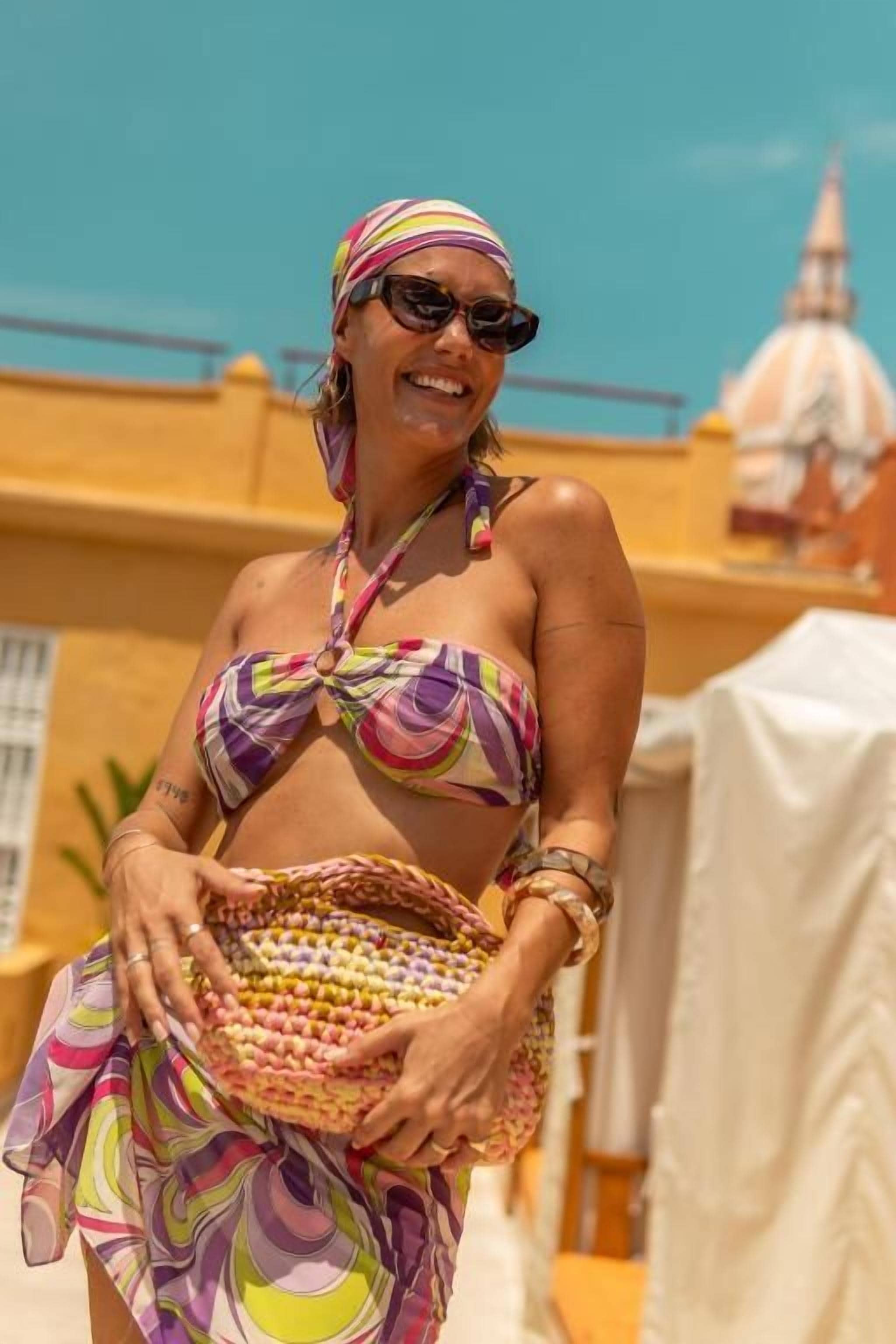 Una persona con traje de baño y diadema coloridos, sosteniendo un bolso tejido, con un edificio y cielo despejado al fondo.