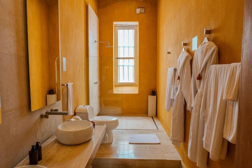 A narrow bathroom with a large mirror, a round sink, a toilet, and a window.
