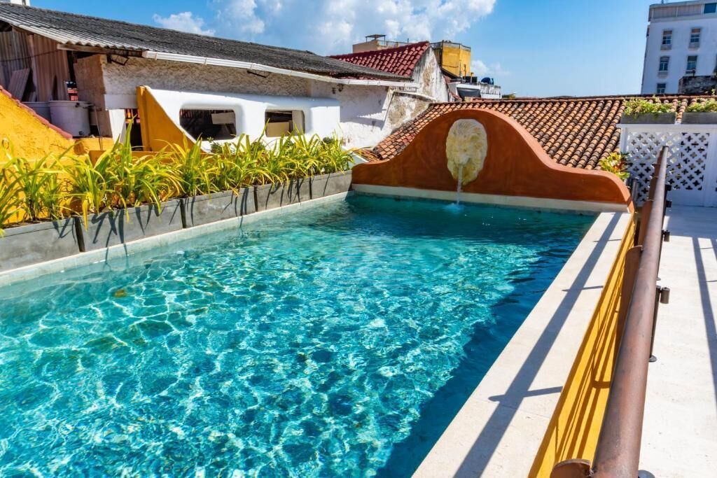 A swimming pool with clear water, surrounded by buildings with red-tiled roofs and a white fence.