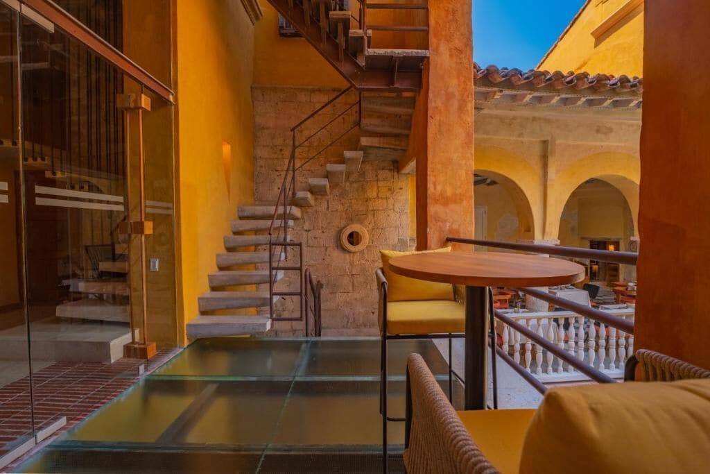 A modern interior with a glass table, yellow chairs, a staircase, and a view of the courtyard.
