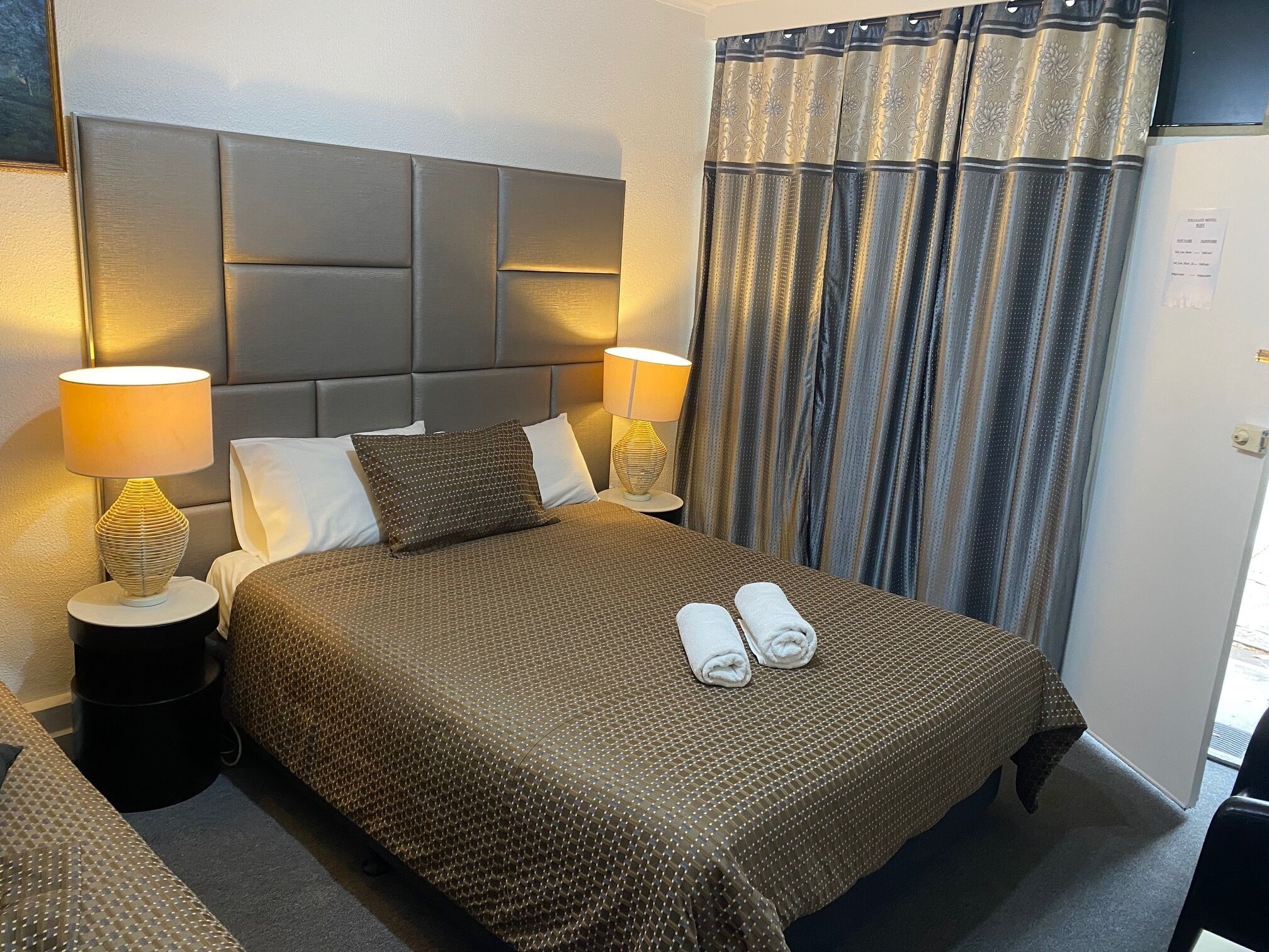 Habitació d'hotel amb un llit gran, dues làmpades laterals, un capçal de llit i una finestra amb cortines.