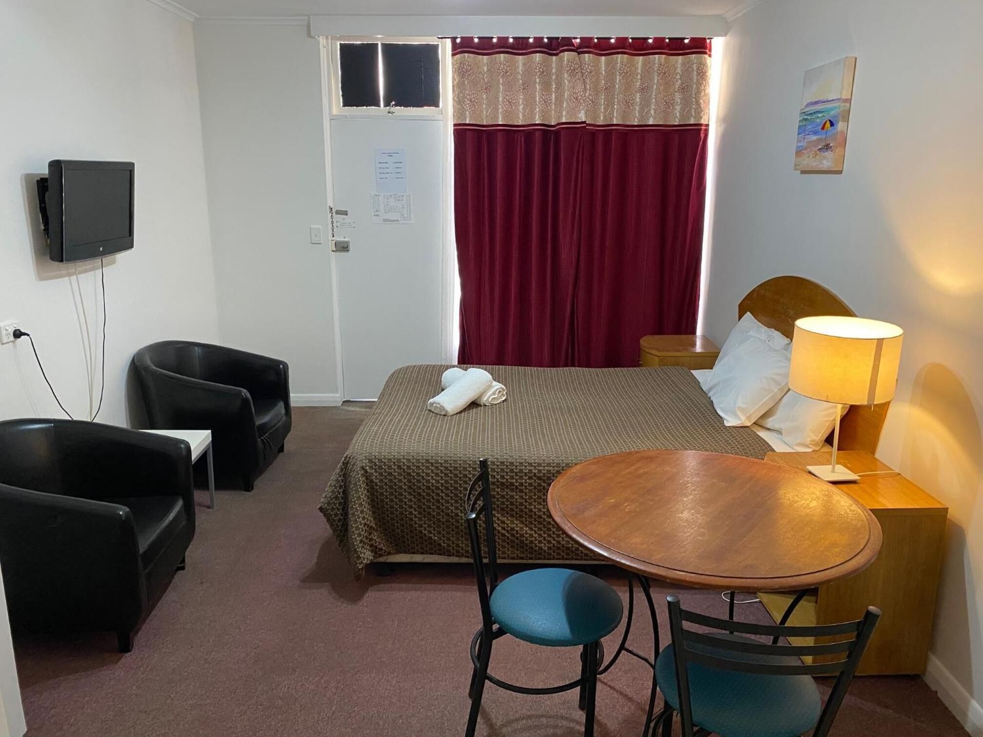 Habitación de hotel con cama, escritorio con lámpara, dos sillas, televisor y ventana con cortinas.