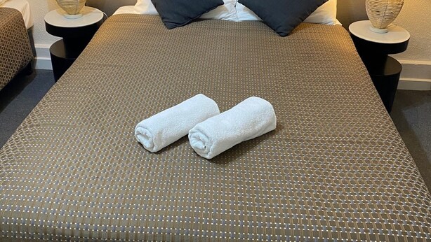 Una cama bien hecha con una colcha marrón texturizada, dos almohadas negras y dos toallas blancas dobladas encima. Hay dos mesitas de noche con lámparas y jarrones decorativos.