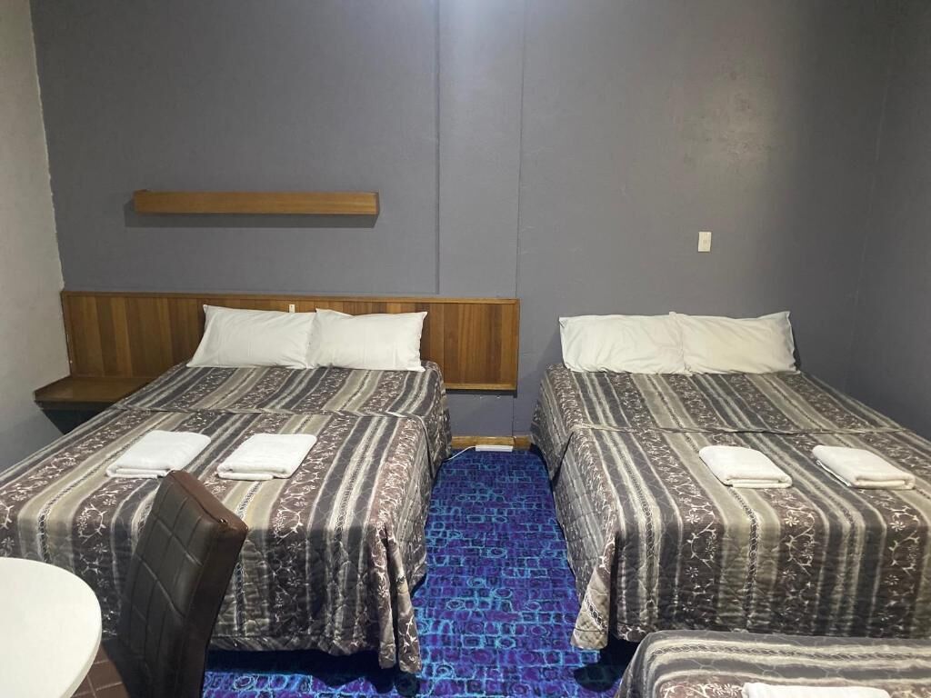 A room with two beds, each with a wooden headboard and white pillows. There is a small table with a chair, and a blue patterned carpet.