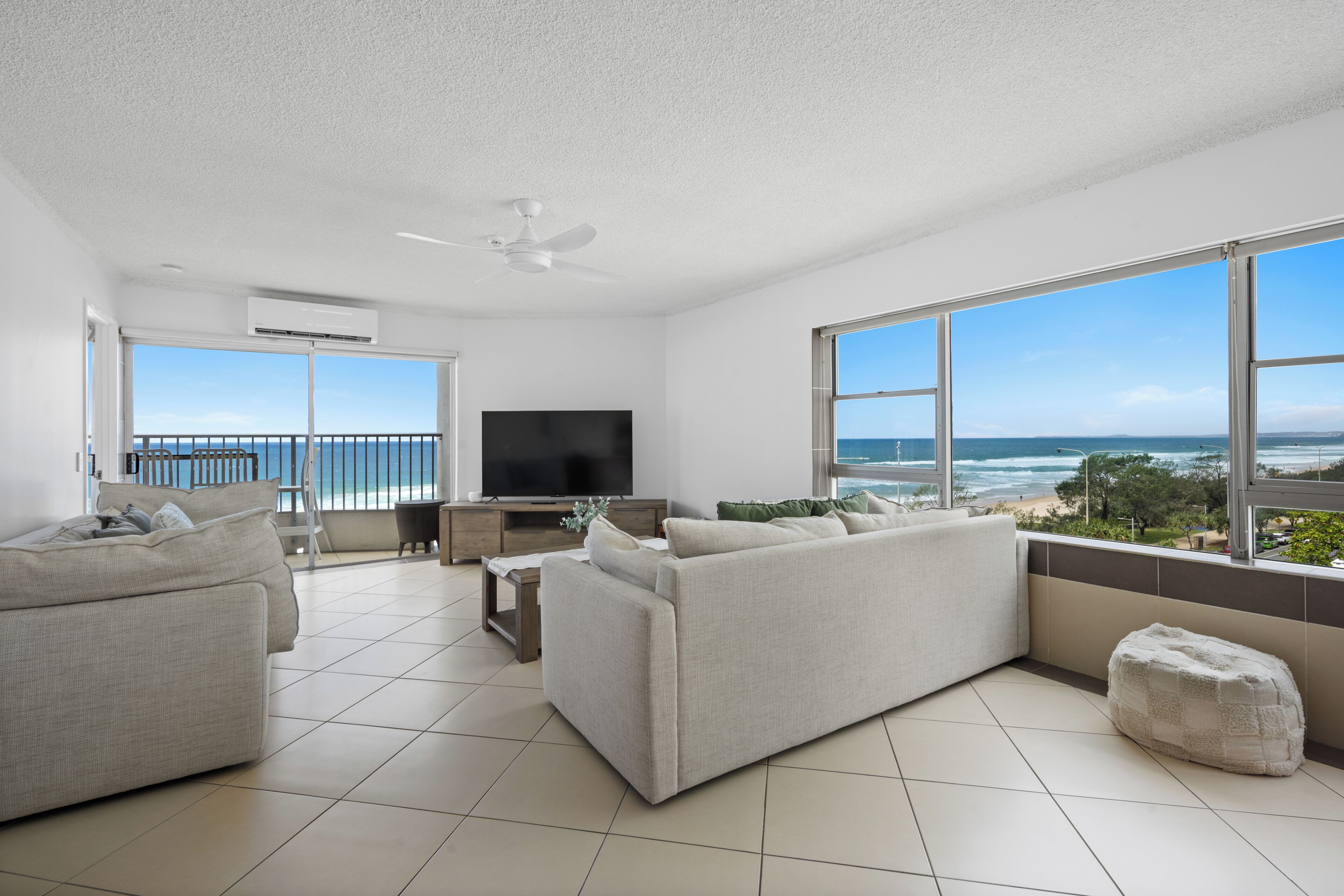 A spacious living room with a large TV, comfortable seating, and a balcony with a view of the ocean.