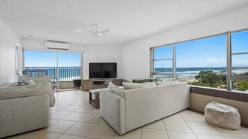 A spacious living room with a large TV, comfortable seating, and a balcony with a view of the ocean.
