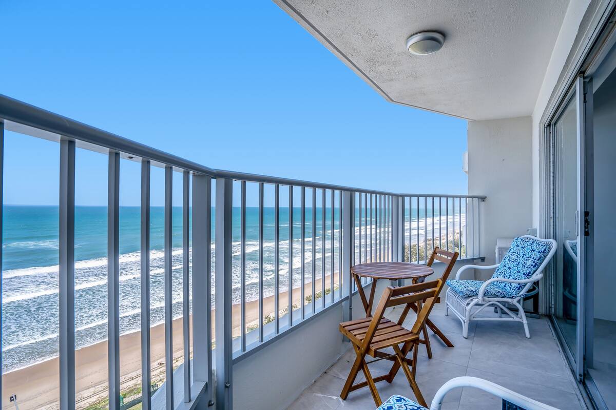 A balcony with a view of the beach and ocean, a table with chairs, and a blue patterned chair.