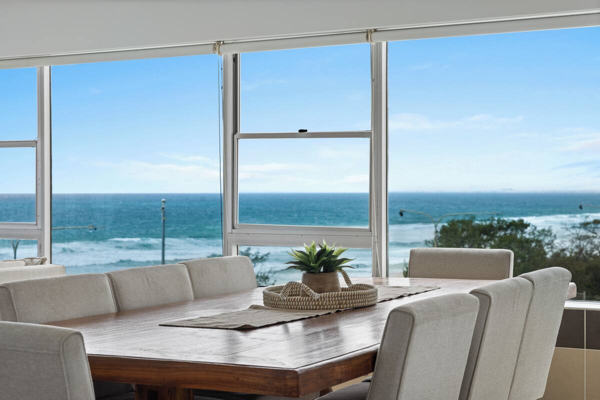A dining area with a wooden table and beige chairs, overlooking the ocean.
