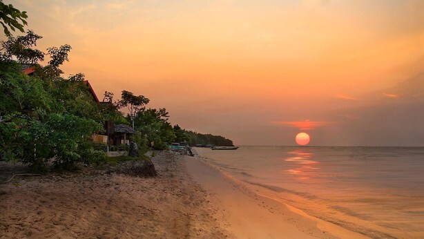 A beach with a sunset, calm sea, and a small house with a thatched roof. - Hotel Santika Palu (Palu)