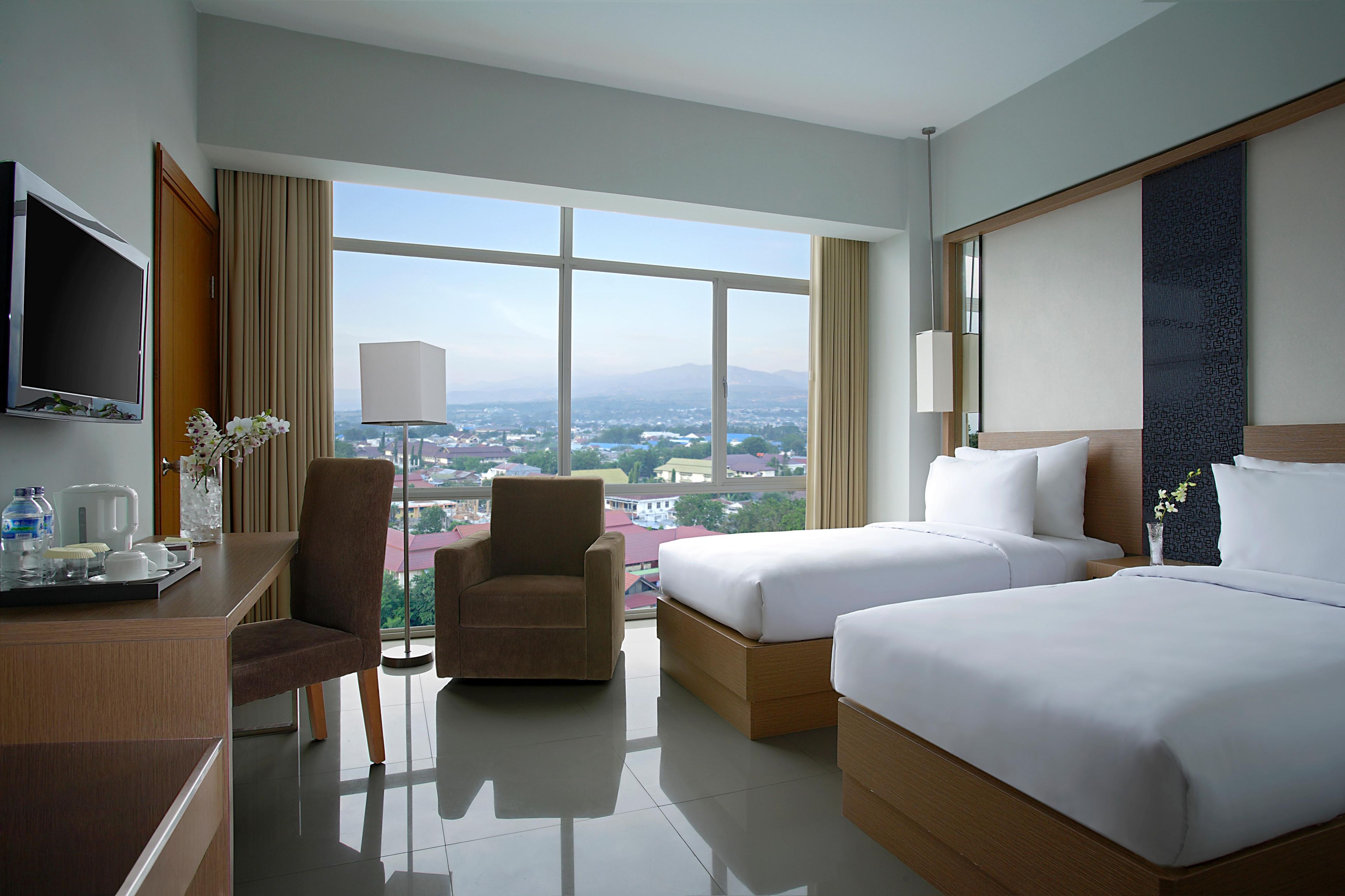 A hotel room with two beds, a desk, a chair, a TV, and a large window with a city view.