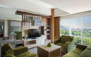 A modern living room with a TV, green sofas, a coffee table with apples, and a city view through large windows.