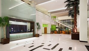 A modern lobby with a reception desk, seating area, staircase, and potted plants. - Hotel Santika Palu (Palu)