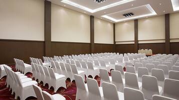 Un salón de conferencias con filas de sillas y mesas blancas, suelo alfombrado e iluminación empotrada en el techo.