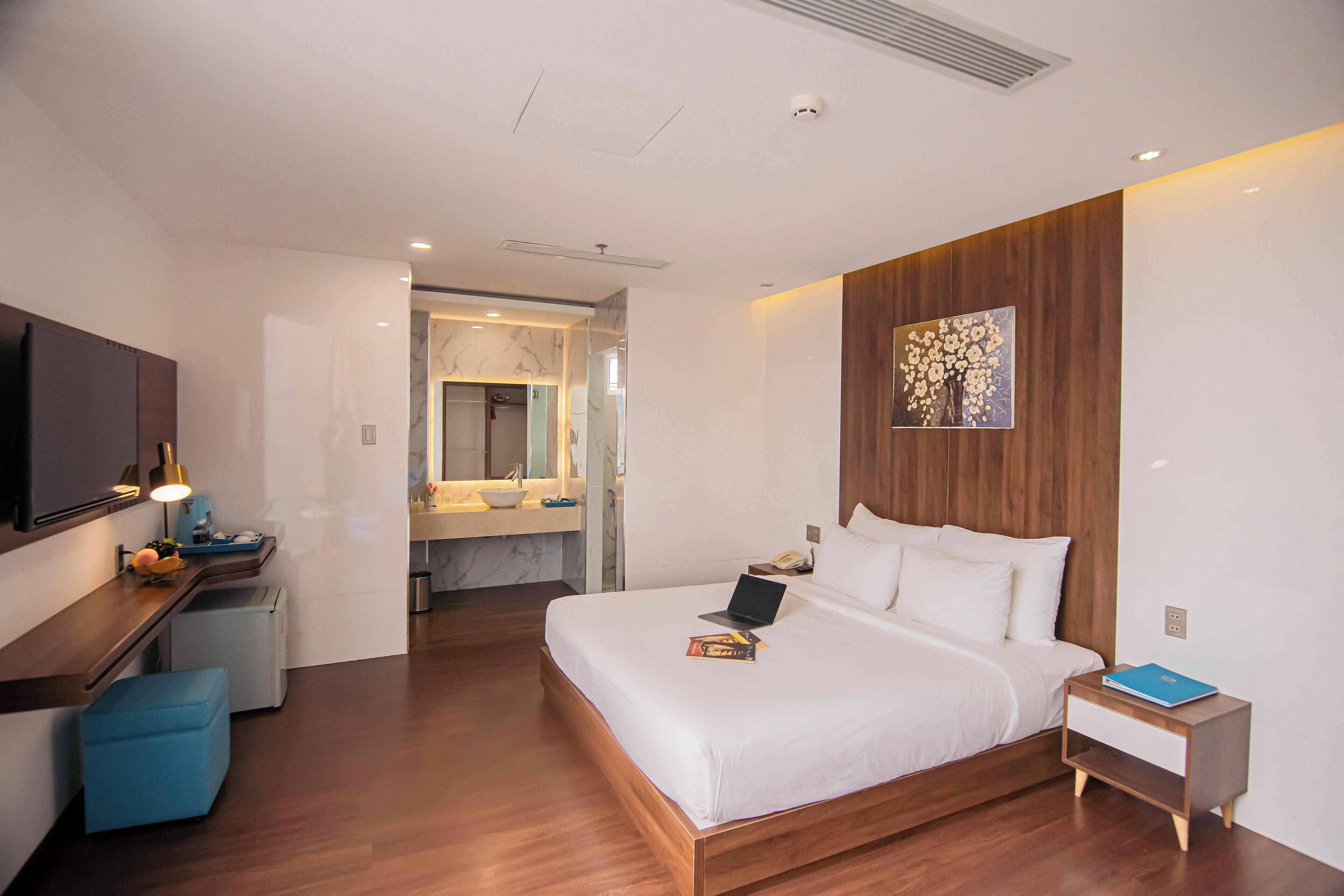 A modern hotel room with a large bed, a desk with a lamp, a small seating area, and a bathroom visible through an open door.