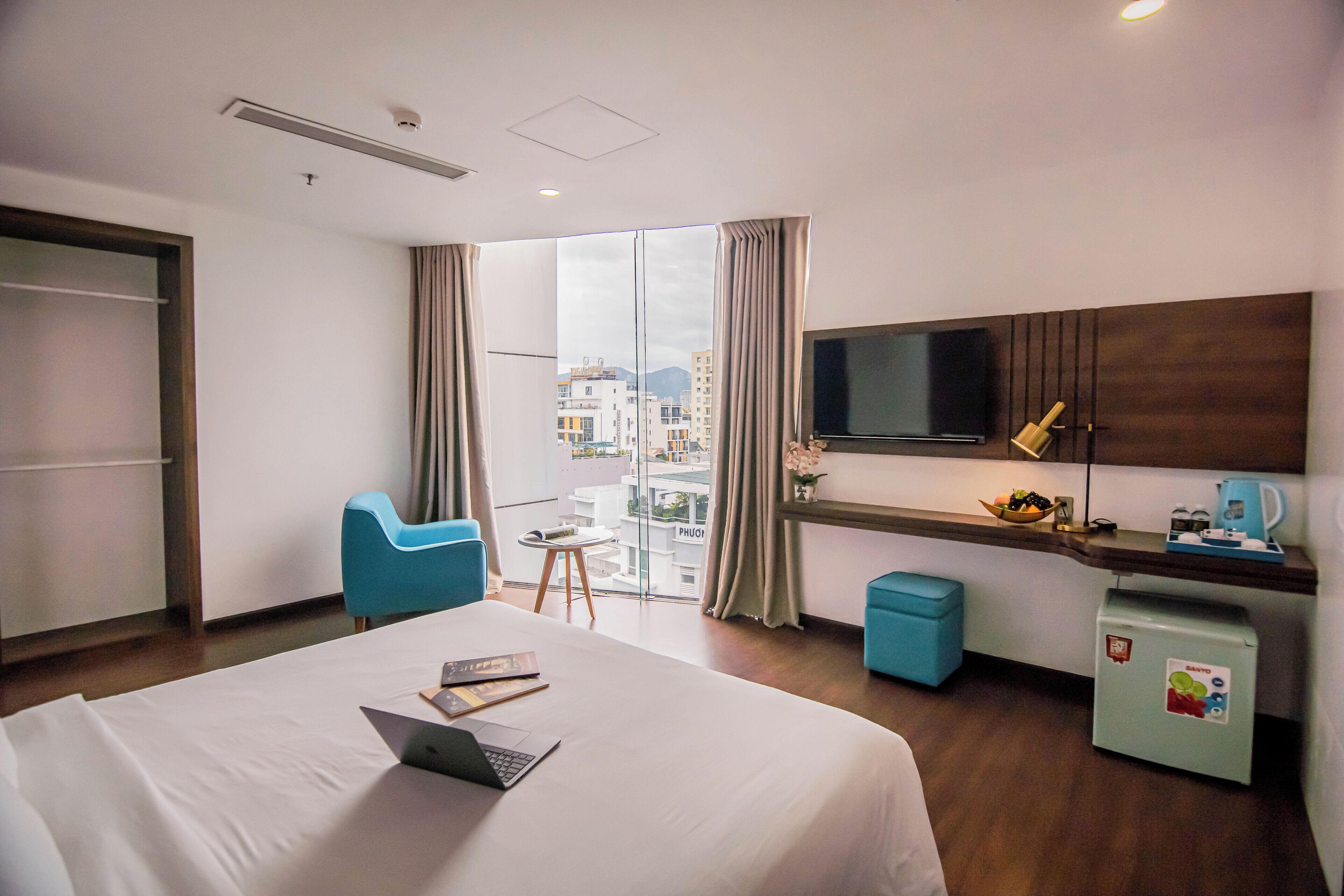 A hotel room with a bed, a desk with a laptop, a TV, and a view of the city.