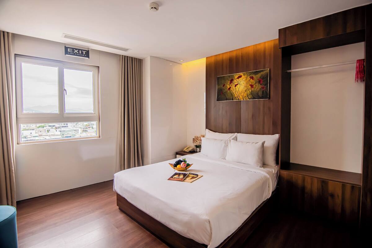 A hotel room with a large bed, a nightstand with a phone, a window with curtains, and a painting on the wall.