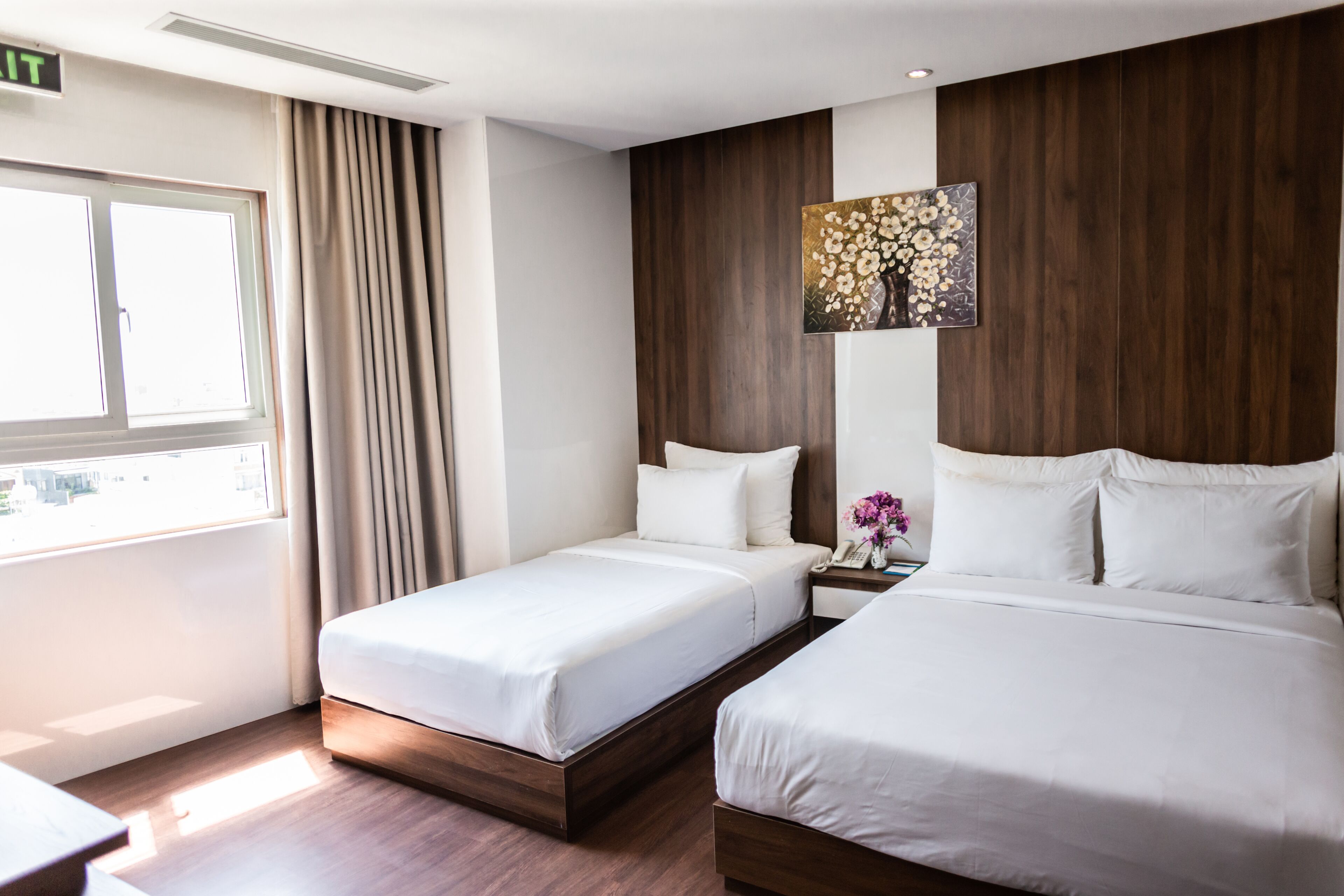 A hotel room with two beds, a nightstand, a painting, and a window with curtains.